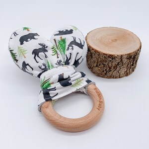 May include: A wooden teething ring with a white fabric loop tied around it. The fabric has a black and green woodland print with bears, moose, and trees. The ring is engraved with the text "Burton Baby".