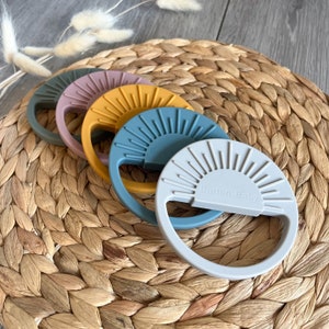 May include: Four silicone teethers in the shape of a sun with a textured surface. The teethers are in the colors gray, blue, yellow, and pink. The teethers have the text "Burton Baby" on them.