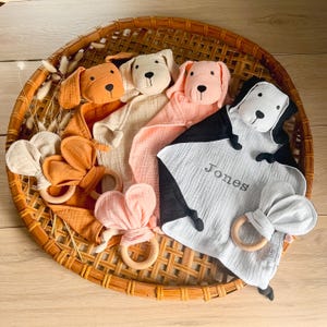 May include: Four plush dog-shaped baby security blankets with attached teethers. The blankets are in beige, pink, orange, and gray. The gray blanket has the name "Jones" embroidered on it.