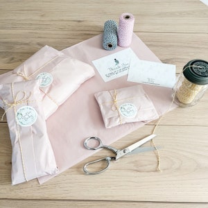 May include: Three small packages wrapped in pink tissue paper with yellow twine and a round sticker that says "The Button Baby" are on a wooden surface.  A pair of silver scissors, two spools of twine, and a thank you card are also on the surface.