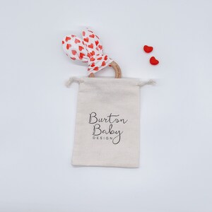 Op de afbeelding: Een witte muslin treksluiting tas met de tekst "Burton Baby Design" erop gedrukt. De tas bevat een houten bijtring met een rode en witte hartjes patroon stof eromheen gebonden. Twee kleine rode hartvormige objecten zijn zichtbaar op de witte achtergrond.