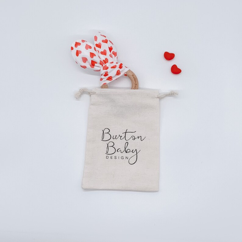 Op de afbeelding: Een witte muslin treksluiting tas met de tekst "Burton Baby Design" erop gedrukt. De tas bevat een houten bijtring met een rode en witte hartjes patroon stof eromheen gebonden. Twee kleine rode hartvormige objecten zijn zichtbaar op de witte achtergrond.