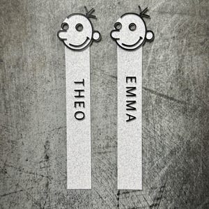 May include: Two personalized bookmarks with a cartoon face design. Each bookmark has a smiling face at the top and a name printed vertically. One says "THEO" and the other says "EMMA".