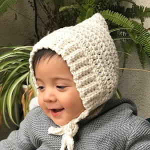 May include: A baby wearing a white crocheted bonnet with a grey knitted sweater.