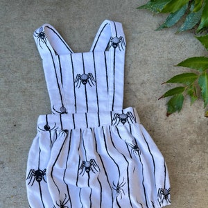 May include: A white and black striped baby onesie with a spider print. The onesie has straps and a ruffled bottom.