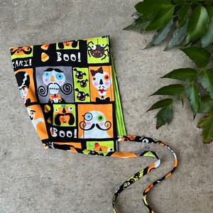 Puede incluir: Un scrunchie de tela de Halloween de color negro, naranja y verde con una variedad de caras de dibujos animados y las palabras "Boo!" y "Scary!" impresas en él.
