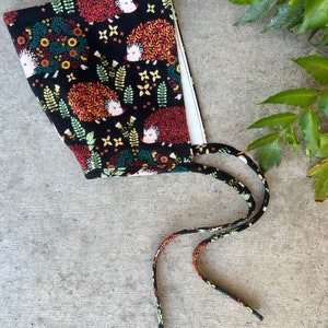 Op de afbeelding: Een zwarte stoffen muts met een witte voering en banden. De stof heeft een kleurrijk patroon van egels, bloemen en bladeren.