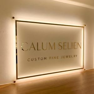 May include: Illuminated sign with the text "CALUM SELIEN CUSTOM FINE JEWELRY" in gold lettering. The sign is rectangular with a gold border and is mounted on a light-colored wall. The sign is lit from behind, creating a soft glow.