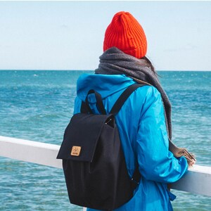 Pu&ograve; includere: Una persona con una giacca blu e un berretto rosso si trova su un molo con vista sull'oceano. Indossa uno zaino in tela nera con una tracolla in pelle marrone.