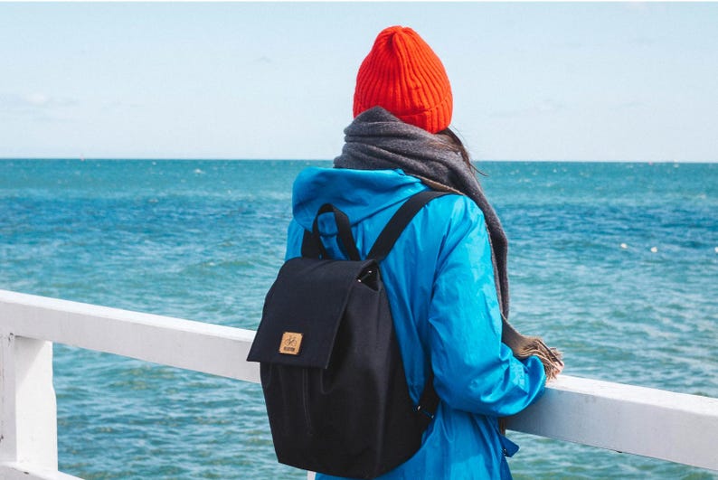 Pu&ograve; includere: Una persona con una giacca blu e un berretto rosso si trova su un molo con vista sull'oceano. Indossa uno zaino in tela nera con una tracolla in pelle marrone.