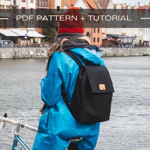 Pu&ograve; includere: Una persona con un impermeabile blu e un cappello rosso &egrave; in piedi vicino a un fiume con uno zaino nero. Lo zaino ha una toppa di cuoio marrone sul davanti. La persona &egrave; in piedi accanto a una bicicletta.