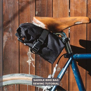 Peut inclure: Un sac de selle noir attaché à un vélo avec une selle en cuir marron. Le sac a une sangle et une boucle noires. Le vélo est appuyé contre une clôture en bois. Le texte "Saddle Bike Bag Sewing Tutorial" est imprimé sous l'image.