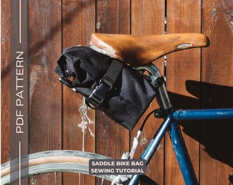 May include: A black saddle bag attached to a bicycle with a brown leather seat. The bag has a black strap and buckle. The bicycle is leaning against a wooden fence. The text "Saddle Bike Bag Sewing Tutorial" is printed below the image.