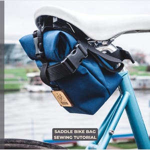 Puede incluir: Una bolsa de sillín azul para bicicleta con una correa y hebilla negras. La bolsa está unida al poste del sillín de la bicicleta. La bolsa tiene una etiqueta de madera con un logotipo de bicicleta. El texto "Saddle Bike Bag Sewing Tutorial" está debajo de la bolsa.