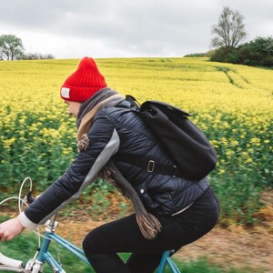 Pu&ograve; includere: Una persona con un berretto rosso e una giacca nera va in bicicletta blu attraverso un campo di fiori gialli.