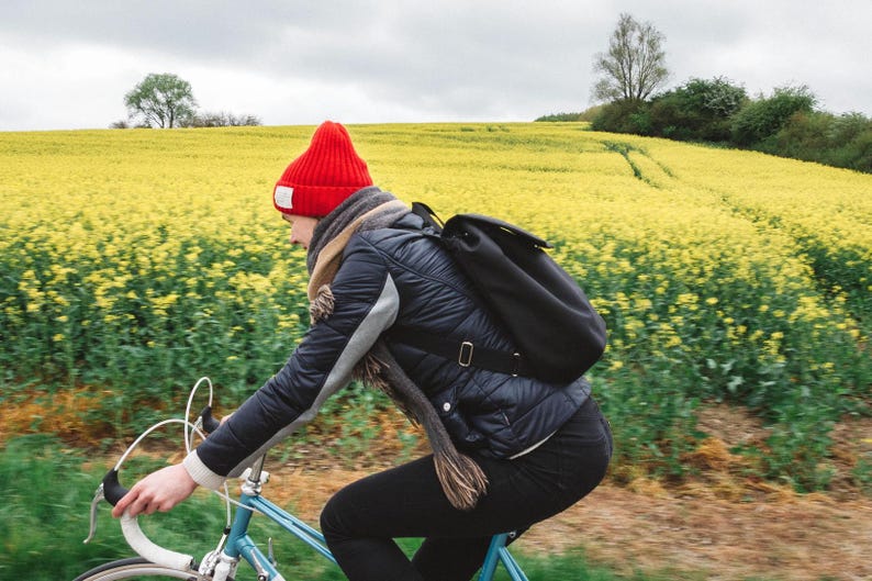 Pu&ograve; includere: Una persona con un berretto rosso e una giacca nera va in bicicletta blu attraverso un campo di fiori gialli.