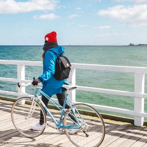 Pu&ograve; includere: Una persona con una giacca blu e un cappello rosso cammina con una bicicletta blu su un molo di legno bianco che si affaccia sul mare.