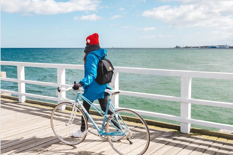 Pu&ograve; includere: Una persona con una giacca blu e un cappello rosso cammina con una bicicletta blu su un molo di legno bianco che si affaccia sul mare.