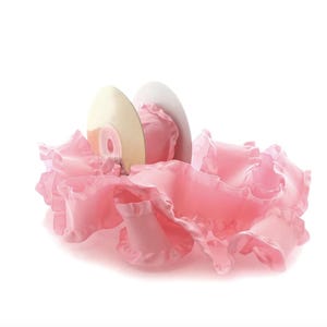 May include: A light pink, ruffled fabric arrangement cradles two off-white discs. The fabric has a delicate, layered appearance, with a soft, flowing texture. The discs are positioned upright within the fabric.