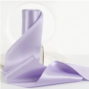 May include: A roll of lavender satin ribbon with a smooth, shiny texture. The ribbon is unspooled, revealing its width and color. The background is a soft white, highlighting the ribbon's delicate hue. Ideal for crafts and gift wrapping.