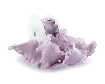 May include: A roll of white ribbon with a lavender ruffled edge. The ribbon is unraveled, with several lavender fabric ruffles scattered around the roll. The ruffles have a soft, textured appearance.