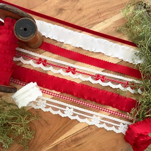 May include: A collection of decorative ribbons including a red ruffled ribbon on a wooden spool, a white scalloped ribbon, a red velvet ribbon, a white lace ribbon with red bows, a red gingham ribbon, and a white lace ribbon with bow motifs.