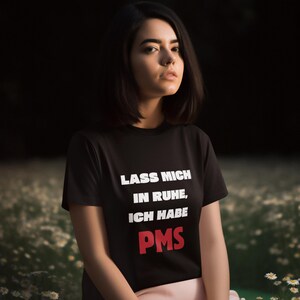 Könnte beinhalten: Eine Frau trägt ein schwarzes T-Shirt mit dem Text "Lass mich in Ruhe, ich habe PMS" in weißer und roter Schrift. Sie sitzt in einem Feld mit weißen Blumen.