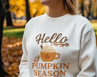 May include: A cream-coloured sweatshirt with the text "Hello Pumpkin Season" in brown script and block letters. The graphic shows a pumpkin spice latte with whipped cream, a cinnamon stick, and autumn leaves. The background is a blurred outdoor scene.