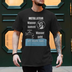 Op de afbeelding: Zwart T-shirt met witte grafische tekst en illustraties. De tekst luidt "INSTALLATEUR Wasser kommt Wasser geht ich bleib cool." De afbeelding bevat een kraan, een loodgieter en waterdruppels. Het shirt heeft korte mouwen.