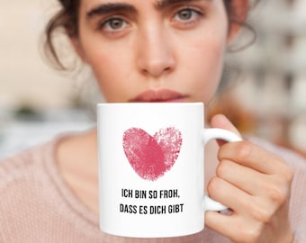 May include: White ceramic mug featuring a red fingerprint heart design. The mug has the German text "ICH BIN SO FROH, DASS ES DICH GIBT" printed below the heart. The mug is held by a person.