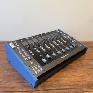 May include: A Solid State Logic audio mixing controller featuring a black control surface with multiple faders, rotary knobs, and illuminated buttons. The unit is encased in a blue frame with white text labels and a navigation pad on the right side.