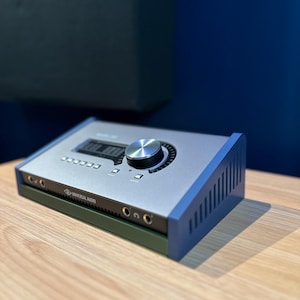 May include: A silver and blue Universal Audio audio interface sits on a wooden surface. The device has a large silver knob, multiple buttons, and the Universal Audio logo. The sides are blue, and the bottom is green.