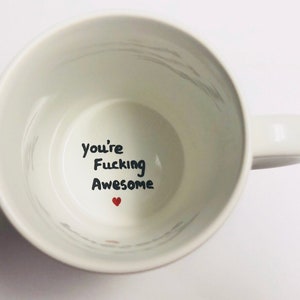May include: White ceramic coffee mug with black handwritten text that reads "You're Fucking Awesome" and a small red heart.