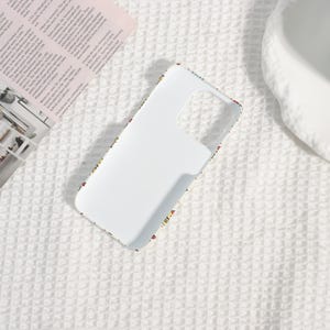 May include: White phone case with a colorful pattern around the edges. The case has cutouts for the camera and buttons. The pattern includes small, colorful shapes and designs. The case is on a white textured surface.