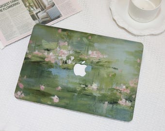 May include: A laptop case featuring an impressionistic painting design in shades of green, blue, and pink. The case has a white Apple logo in the center. The case is on a white surface with a newspaper and a white teacup.