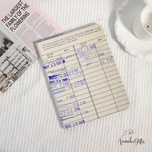 May include: A vintage-style library card notebook with blue stamped dates and a cream-colored background. The dates include October 18, 1989, and August 19, 1989. A pink magazine with the text "THE LARGEST FAMILY OF THE FLOWERING" is in the background.