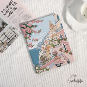 Könnte beinhalten: Ein Notizbuch mit einer farbenfrohen Illustration einer italienischen Küstenstadt, mit Gebäuden, einem Roller und rosa Blumen. Der Einband hat einen hellblauen Himmelshintergrund und ein Blumenmuster. Der Text "THE LARGEST FAMILY OF THE FLOWERING" ist sichtbar.