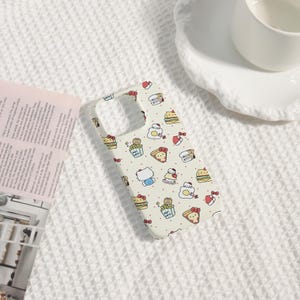 May include: A cream-colored phone case featuring a repeating pattern of Hello Kitty characters and food items like cake, popcorn, and eggs. The case has a cutout for the camera and buttons. Small red polka dots are scattered throughout the design.