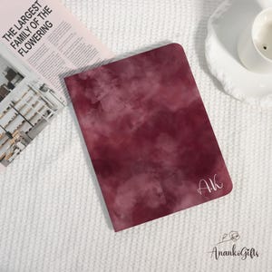 Könnte beinhalten: Ein weinrotes Notizbuch mit aquarellartigem Design und den Initialen "AK" in weißer Schrift. Das Notizbuch liegt auf einer weißen, strukturierten Oberfläche, neben einer Zeitschrift und einem weißen Teller mit einer Tasse.