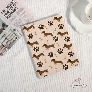 May include: A beige tablet case featuring a repeating pattern of brown dachshunds, paw prints, and bones. The case is on a white surface with a newspaper and a teacup in the background. The text on the newspaper reads "THE LARGEST FAMILY OF THE FLOWERING".