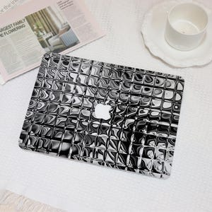 May include: A laptop computer featuring a black and white grid pattern with a distorted, liquid-like effect across the surface. The center of the lid displays a white Apple logo, contrasting against the monochromatic, wavy, and reflective square tiles.