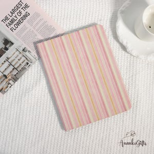 May include: A pink notebook with vertical stripes in light pink and yellow. The notebook is open, and a magazine with the text "THE LARGEST FAMILY OF THE FLOWERING" is visible. A white teacup and saucer are in the background.