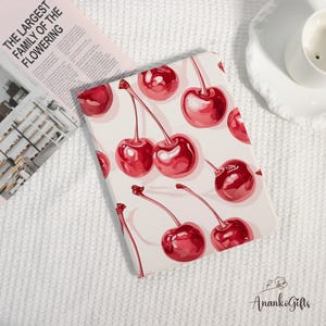 May include: A rectangular notebook with a white background and a pattern of red cherries. The notebook is open, and the text "THE LARGEST FAMILY OF THE FLOWERING" is visible on a pink newspaper. The notebook is on a white textured surface.