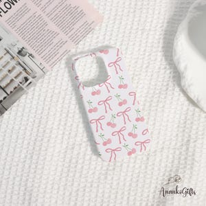 May include: White phone case with a repeating pattern of pink cherries and bows. The case has a cutout for the camera and buttons. The design is feminine and playful, perfect for adding a touch of personality to a phone.
