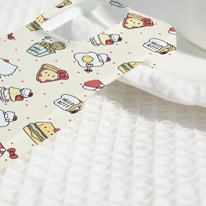 May include: A light-colored phone case featuring a repeating pattern of Hello Kitty characters and food items like pizza, popcorn, and sandwiches. The case has a smooth finish and a white border. Small red hearts are scattered throughout the design.