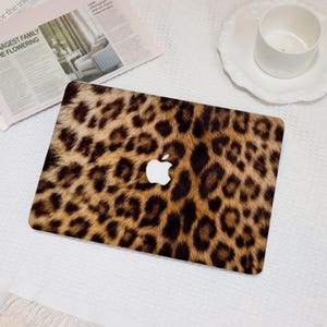 May include: A laptop skin featuring a leopard print design in shades of brown and black. The Apple logo is visible in the center. The skin is placed on a white surface with a newspaper and a cup.