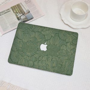 May include: Green laptop case featuring a floral design and the Apple logo. The case has a textured pattern of stylized flowers in green and gold. The case is on a white surface with a newspaper and a white teacup.
