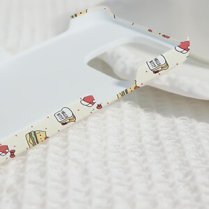 May include: A white phone case with a patterned edge featuring Hello Kitty, sandwiches, and Santa hats. The case has a cutout for the camera and is set against a white, textured background.