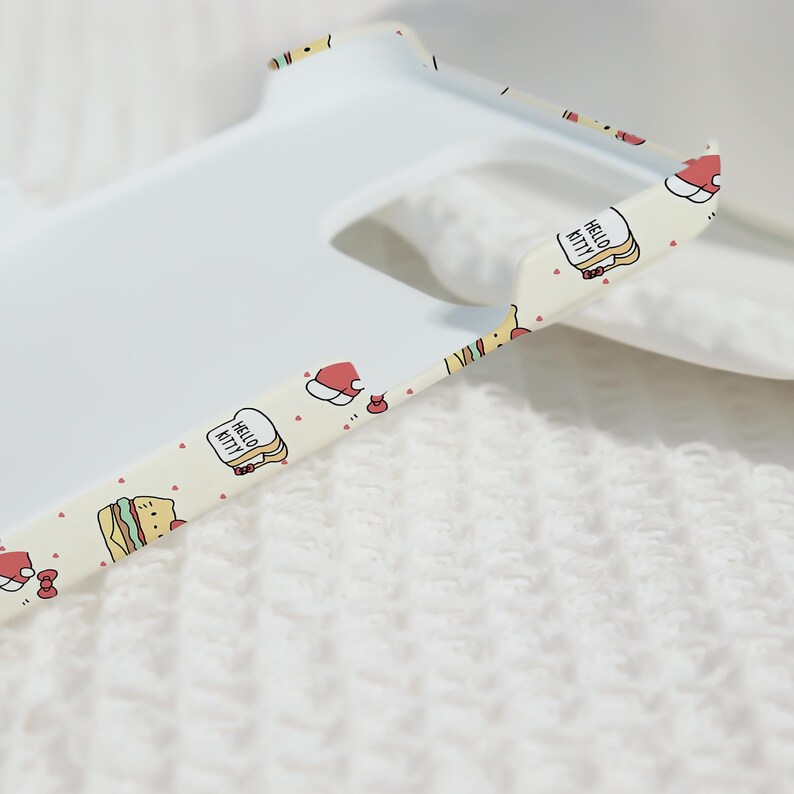 May include: A white phone case with a patterned edge featuring Hello Kitty, sandwiches, and Santa hats. The case has a cutout for the camera and is set against a white, textured background.