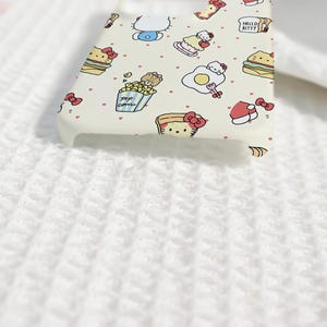 May include: A white phone case featuring a repeating pattern of Hello Kitty and food-themed illustrations. The case includes images of sandwiches, popcorn, and eggs, with the words "Hello Kitty" printed on the side.